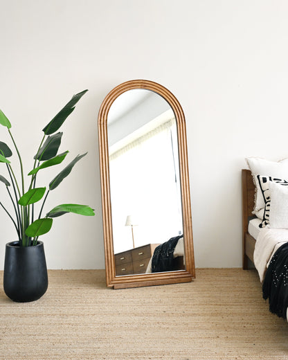 Wooden floor mirror leaning against a white wall in a bedroom with a plant and bed.