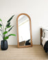 Wooden floor mirror leaning against a white wall in a bedroom with a plant and bed.