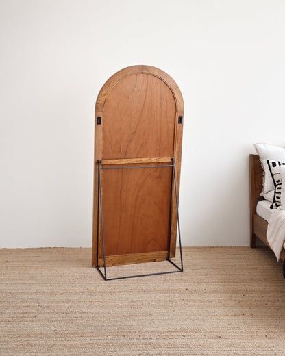Wooden standing mirror with curved top in a room with a bed and white wall.