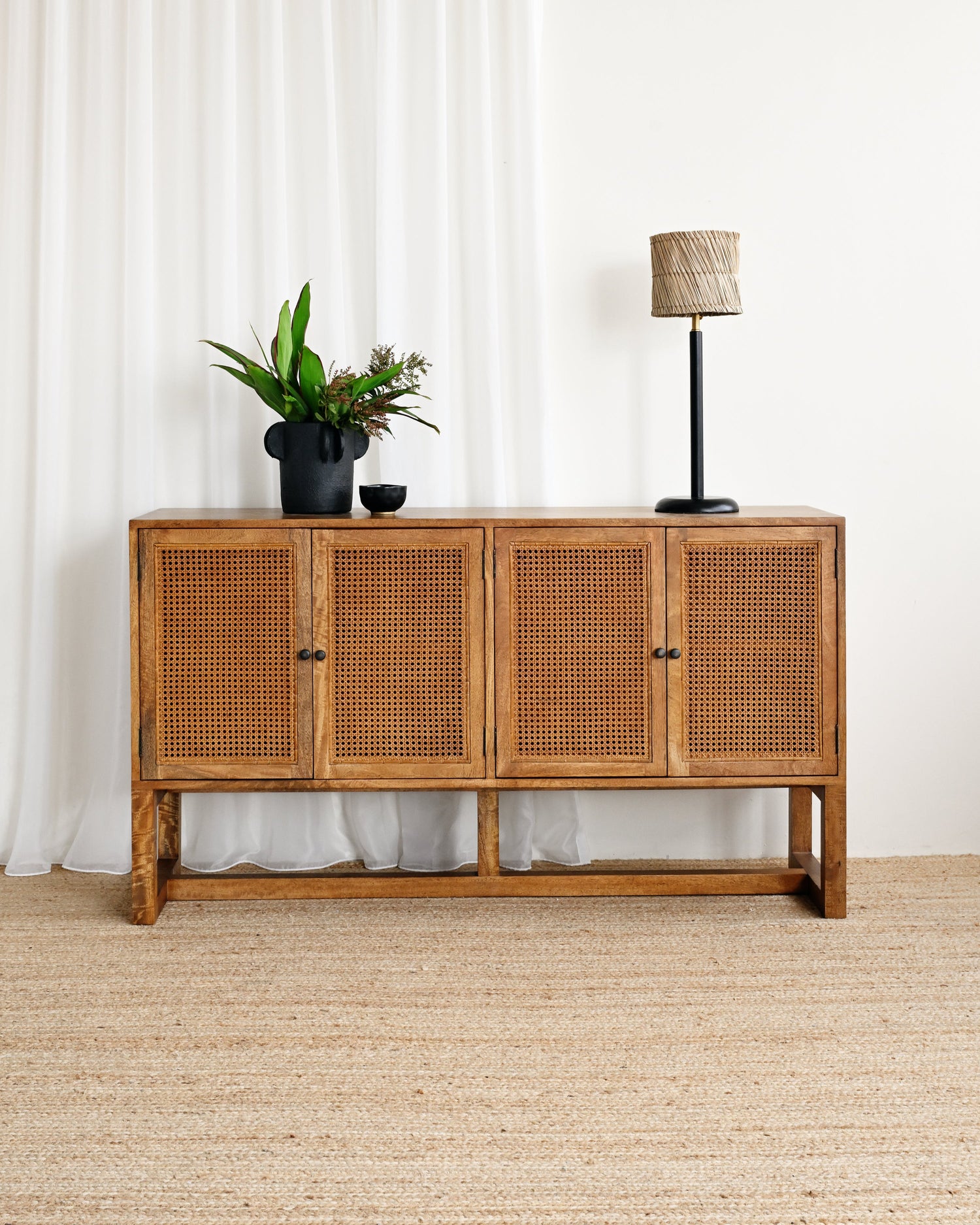 Wooden sideboard with rattan doors in a room with white curtains and a lamp.