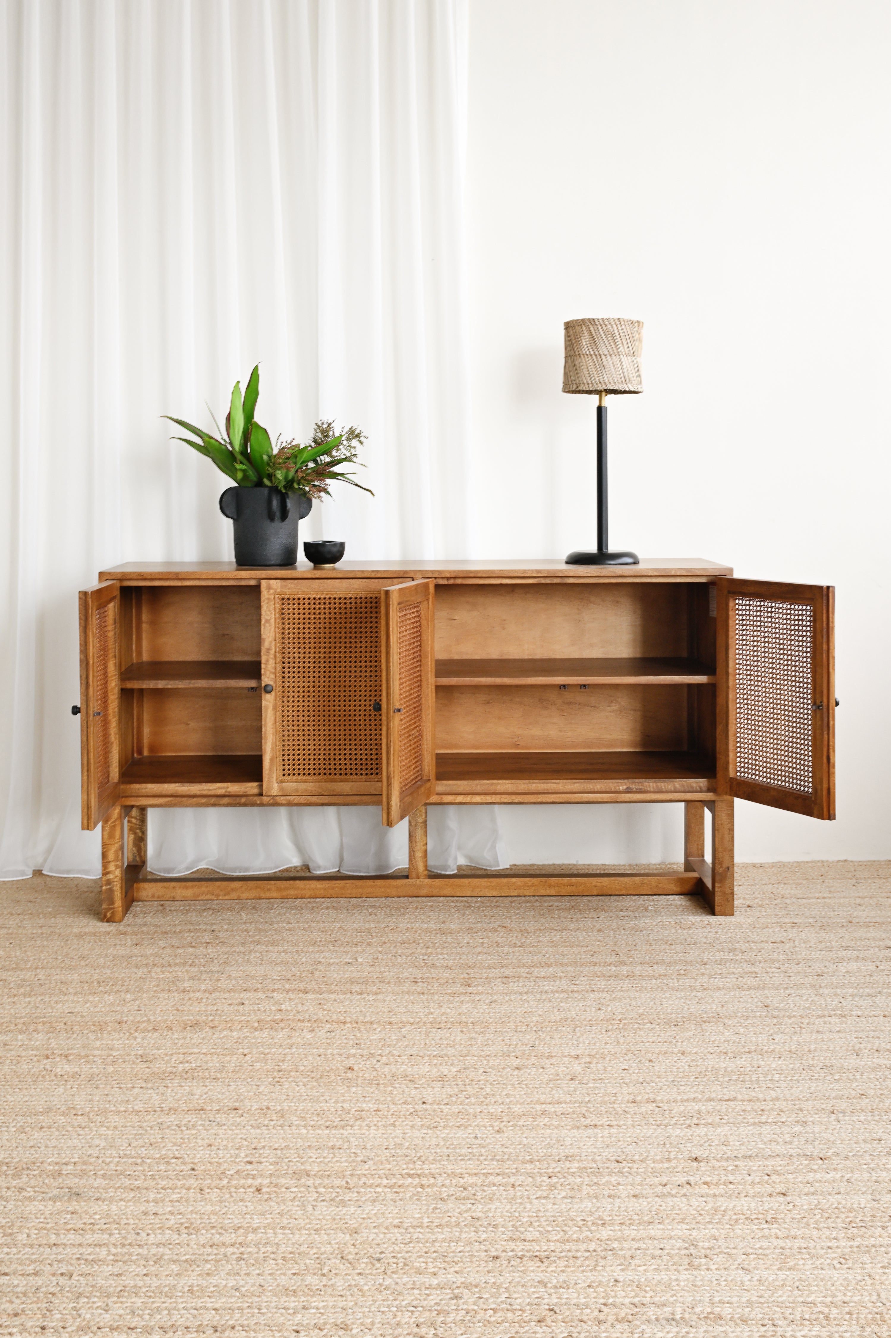 Wooden sideboard with open doors, potted plant, and lamp against a white curtain background.