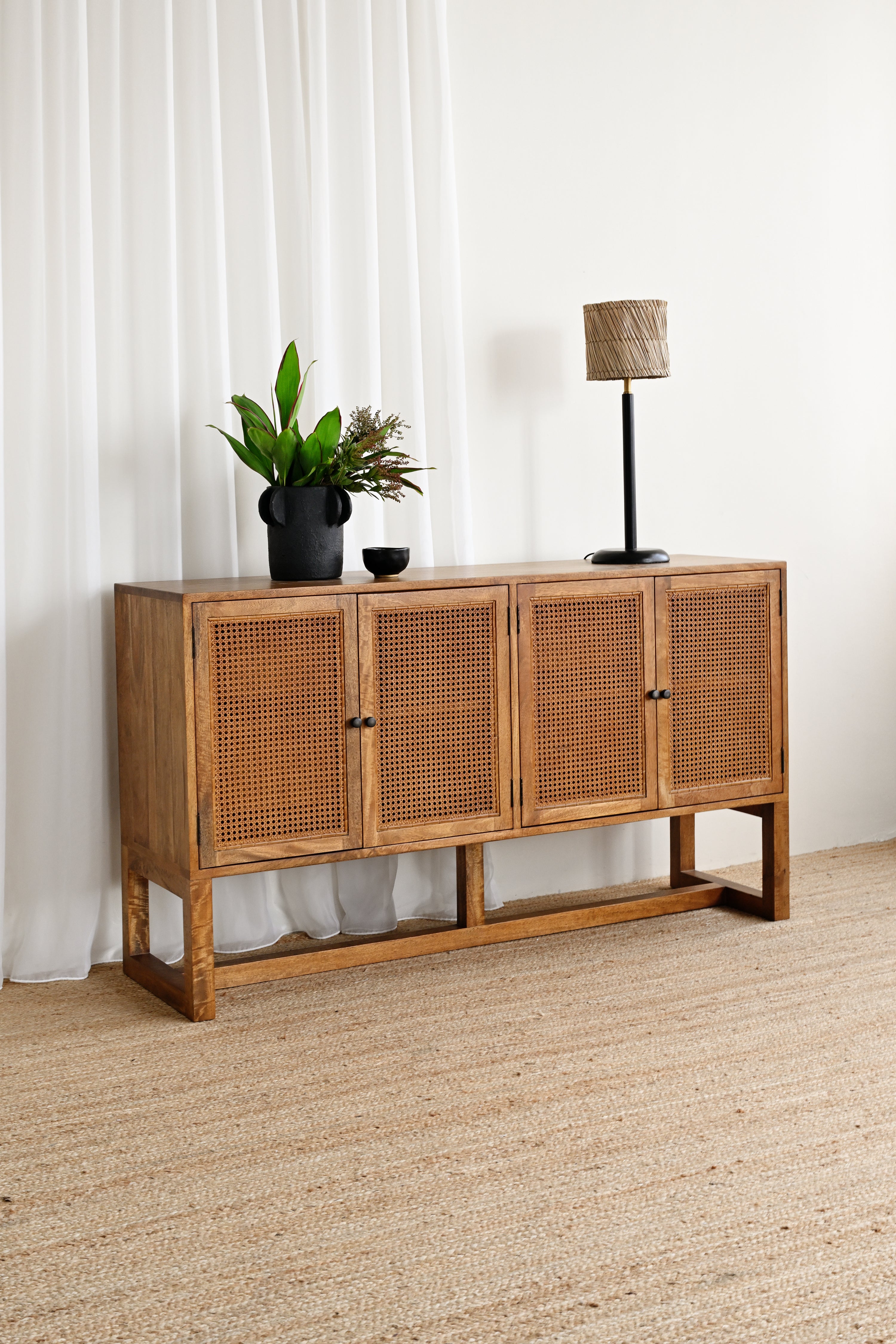 Wooden sideboard with rattan doors in a room with white curtains and a lamp.