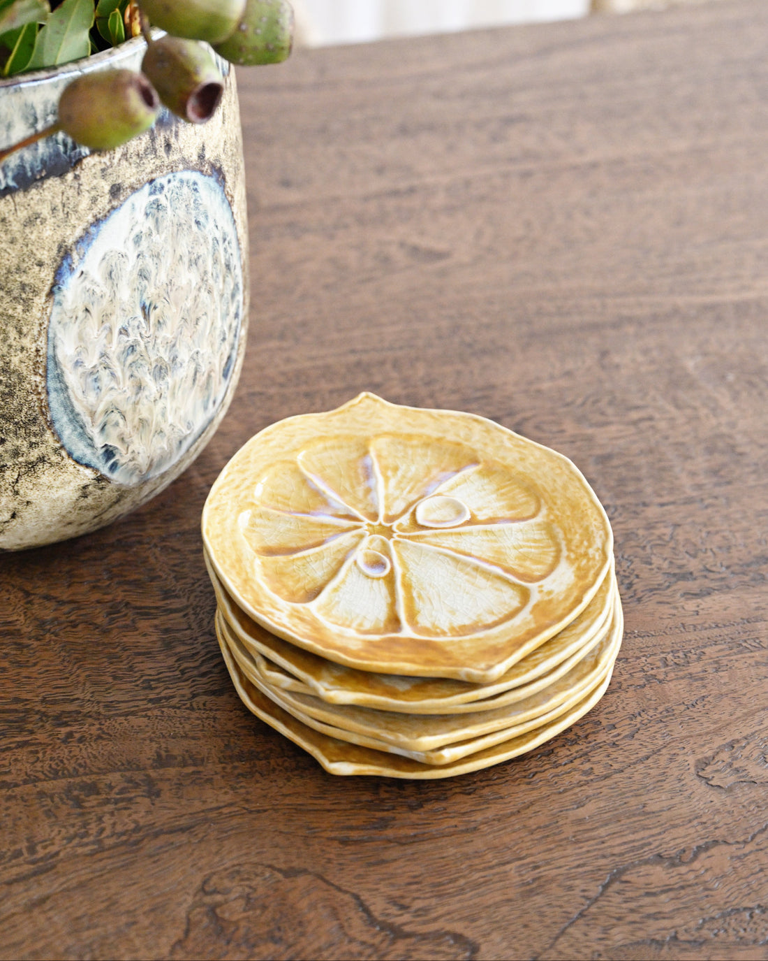 Stack of pancakes with lemon slices on a wooden table next to a vase with greenery