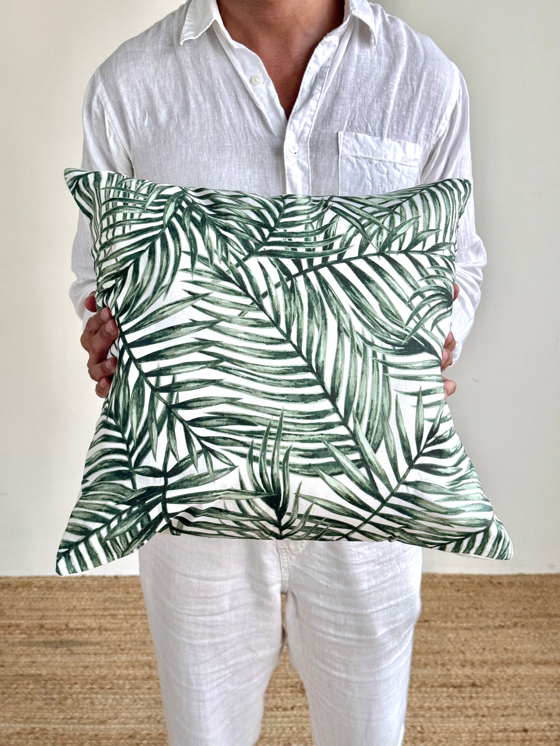 A person wearing a white linen shirt and white pants holds a square throw pillow with a green palm leaf pattern on a white background.