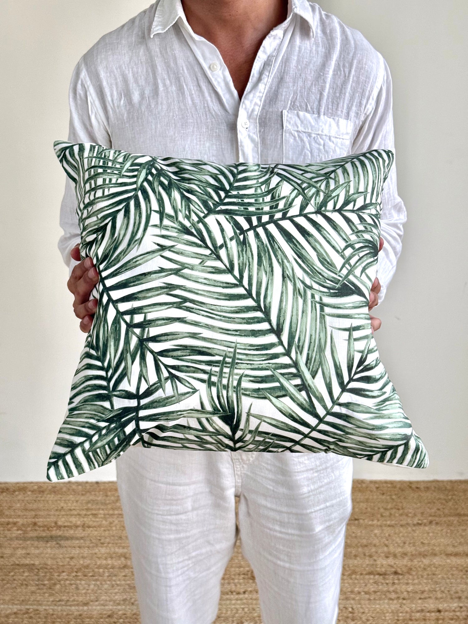 A person wearing a white linen shirt and white pants holds a square throw pillow with a green palm leaf pattern on a white background.