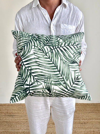 A person wearing a white linen shirt and white pants holds a square throw pillow with a green palm leaf pattern on a white background.