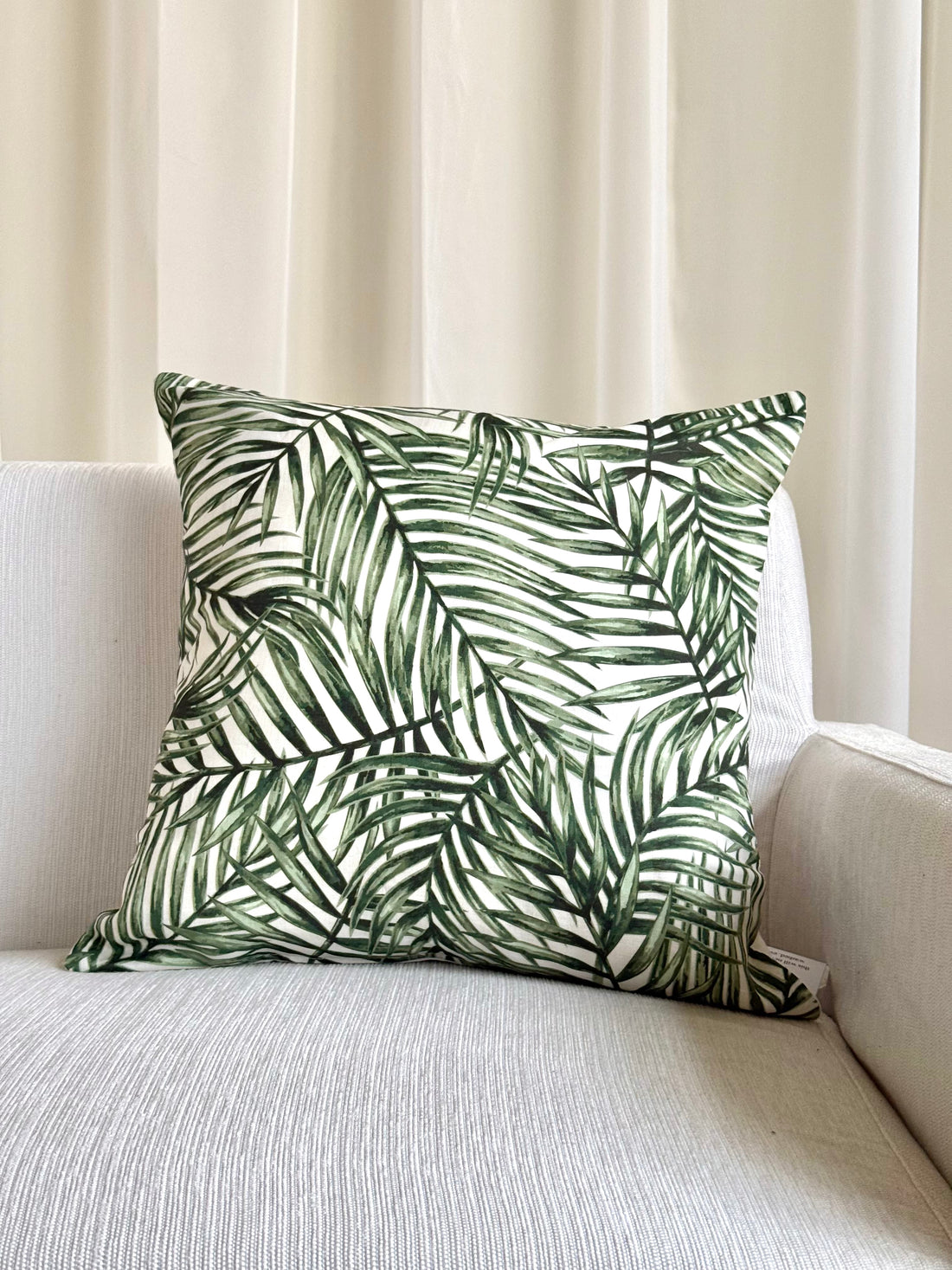 A square throw pillow with a green palm leaf pattern on a white background, resting on a light beige textured sofa in front of cream-colored curtains.