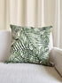 A square throw pillow with a green palm leaf pattern on a white background, resting on a light beige textured sofa in front of cream-colored curtains.