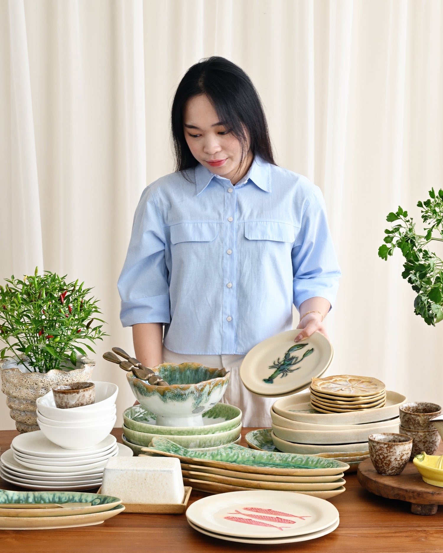 Woman arranging ceramic plates and bowls on a wooden table with plants in the background.