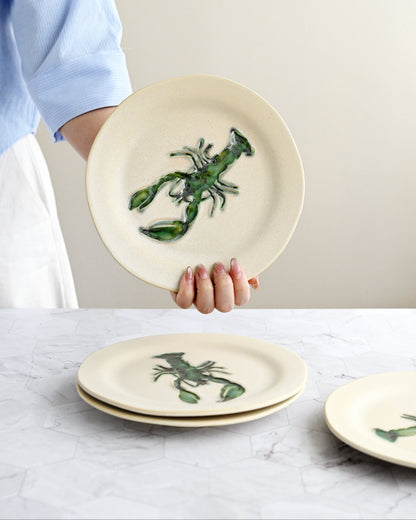 Person holding a ceramic plate with a green lobster design on a marble surface.