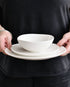 Person holding a white bowl and plate set against a neutral background