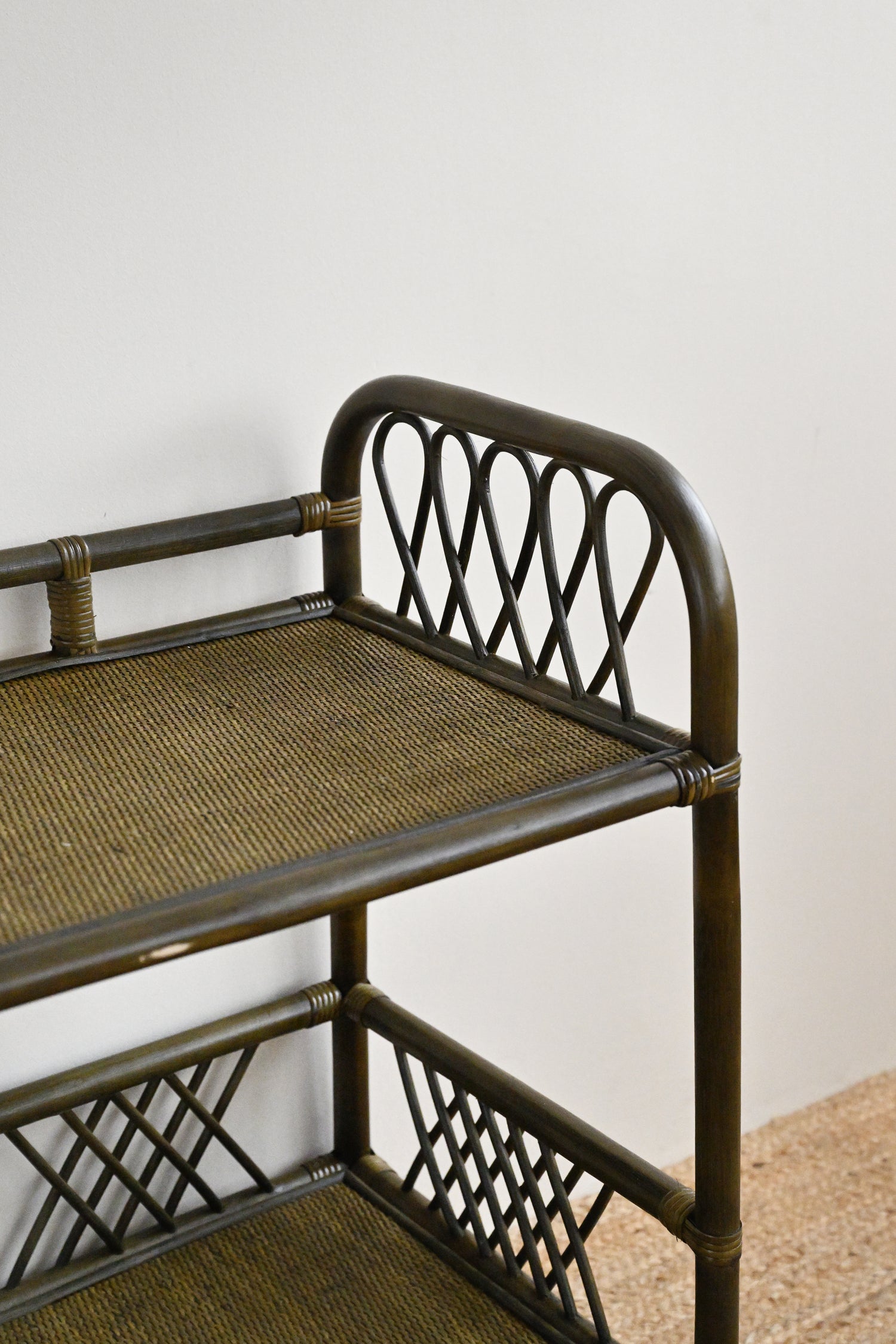 Rattan bar cart with woven seat and shelves against a white wall.