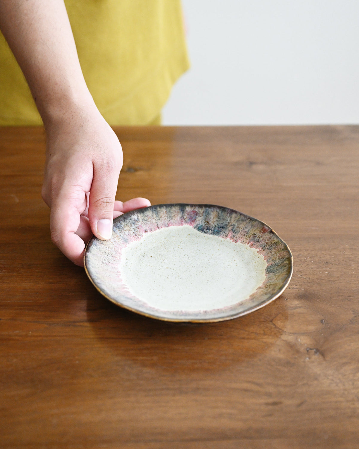 A hand holding a small round ceramic plate with a speckled off-white center and a mottled blue, purple, and gold rim, set on a wooden table.