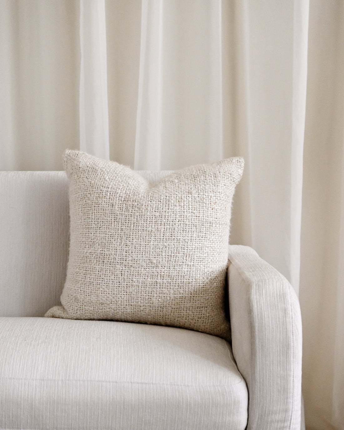 A square beige textured throw pillow resting on a light beige upholstered sofa in front of sheer cream curtains.