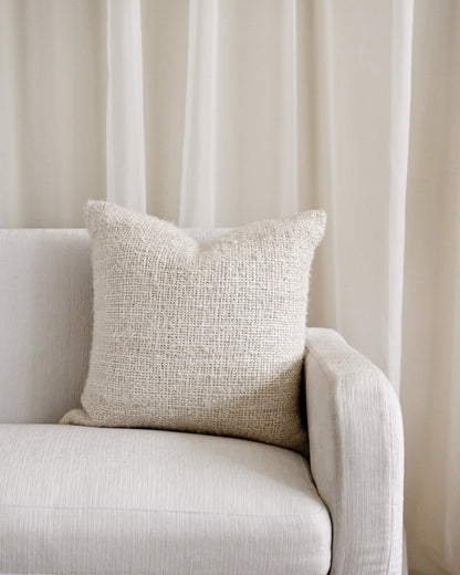 A square beige textured throw pillow resting on a light beige upholstered sofa in front of sheer cream curtains.