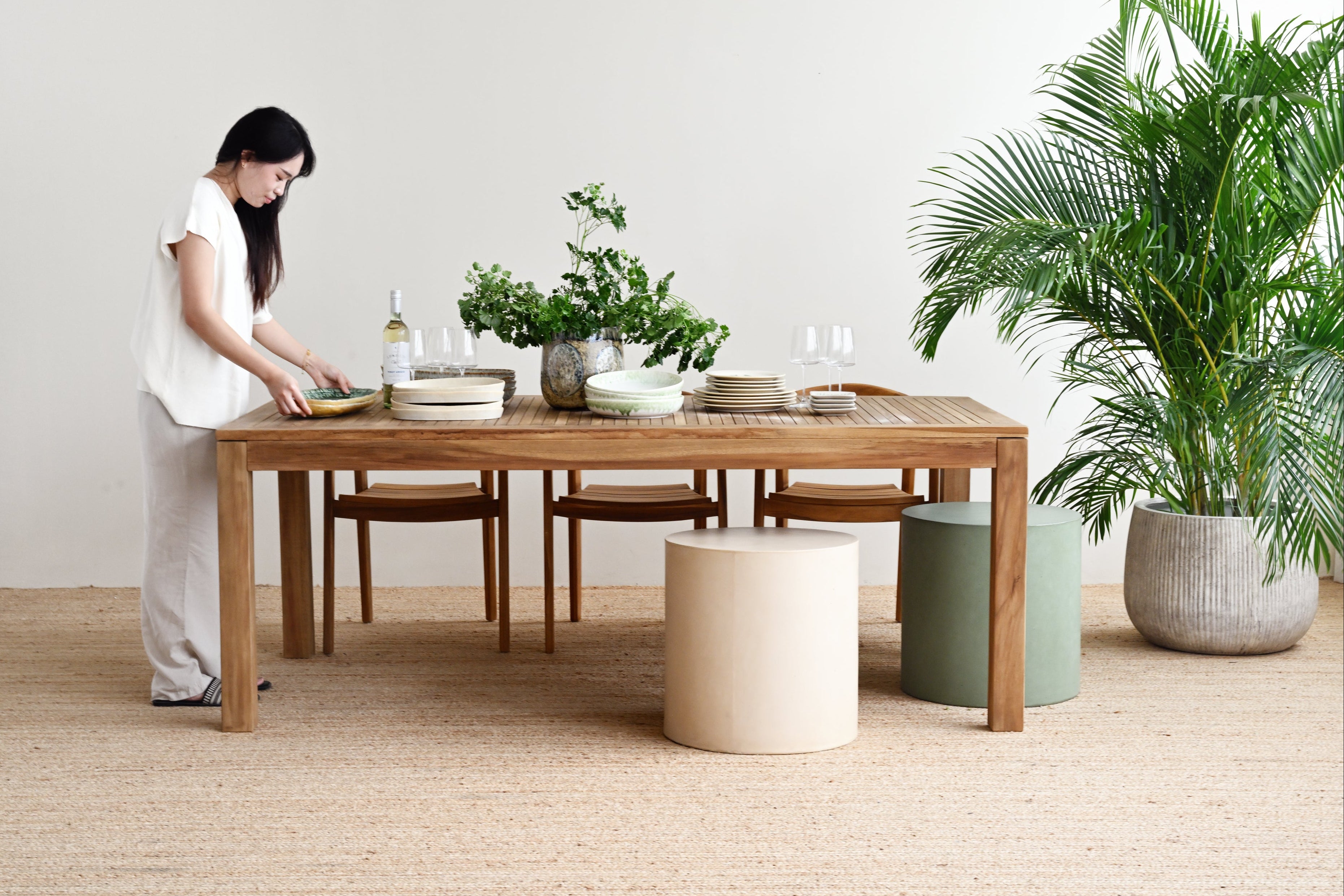 Woman arranging items on a wooden dining table in a minimalistic room with plants.