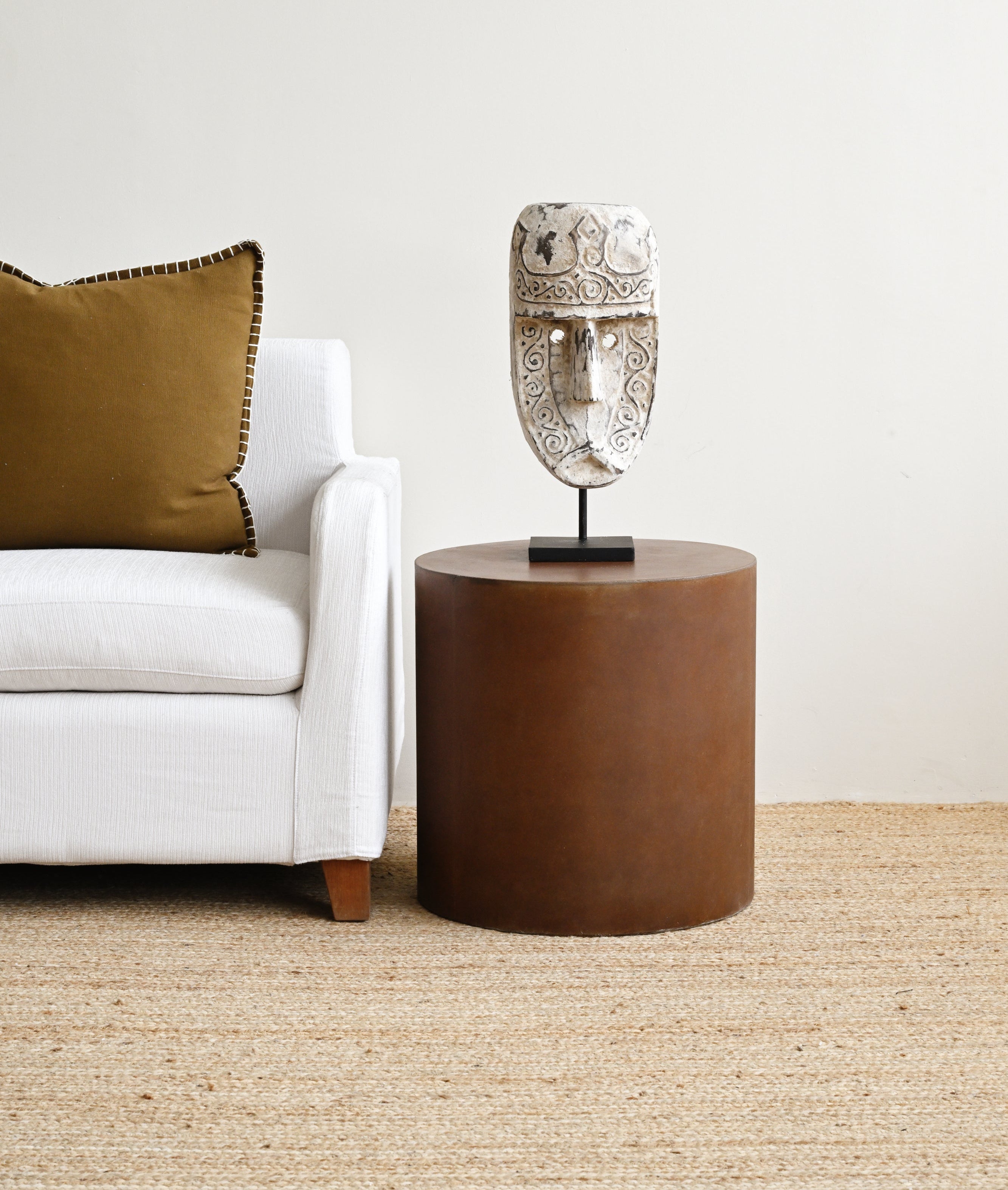 Cylindrical brown side table with a decorative mask on top next to a white sofa with a green pillow.