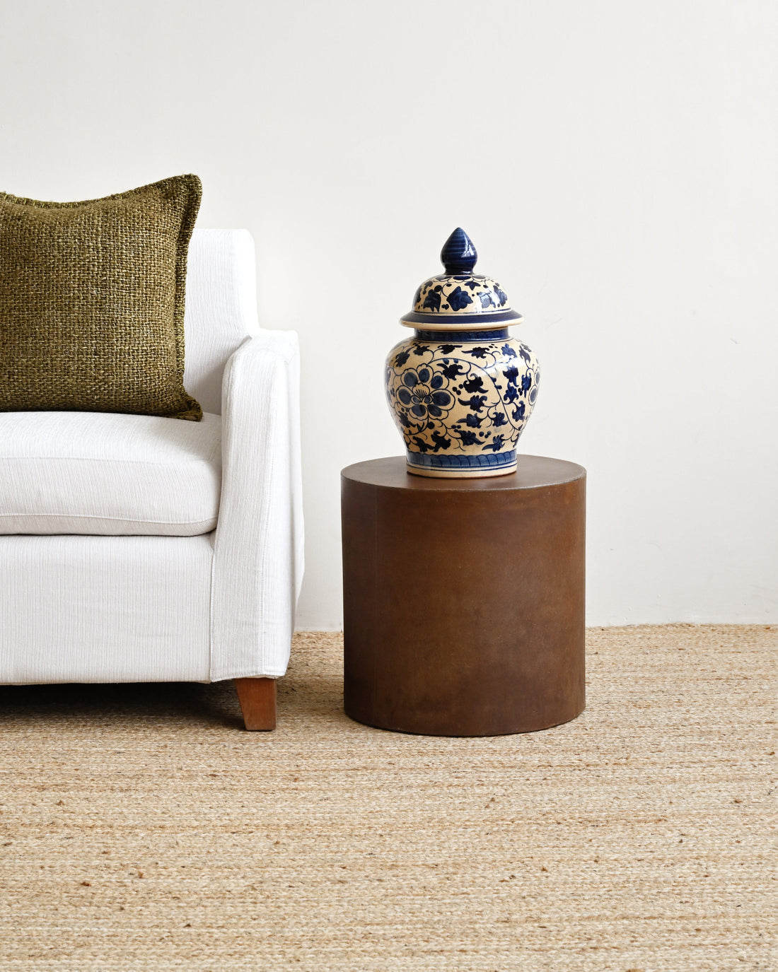 Vase on a small round wooden table next to a white sofa with a green pillow.