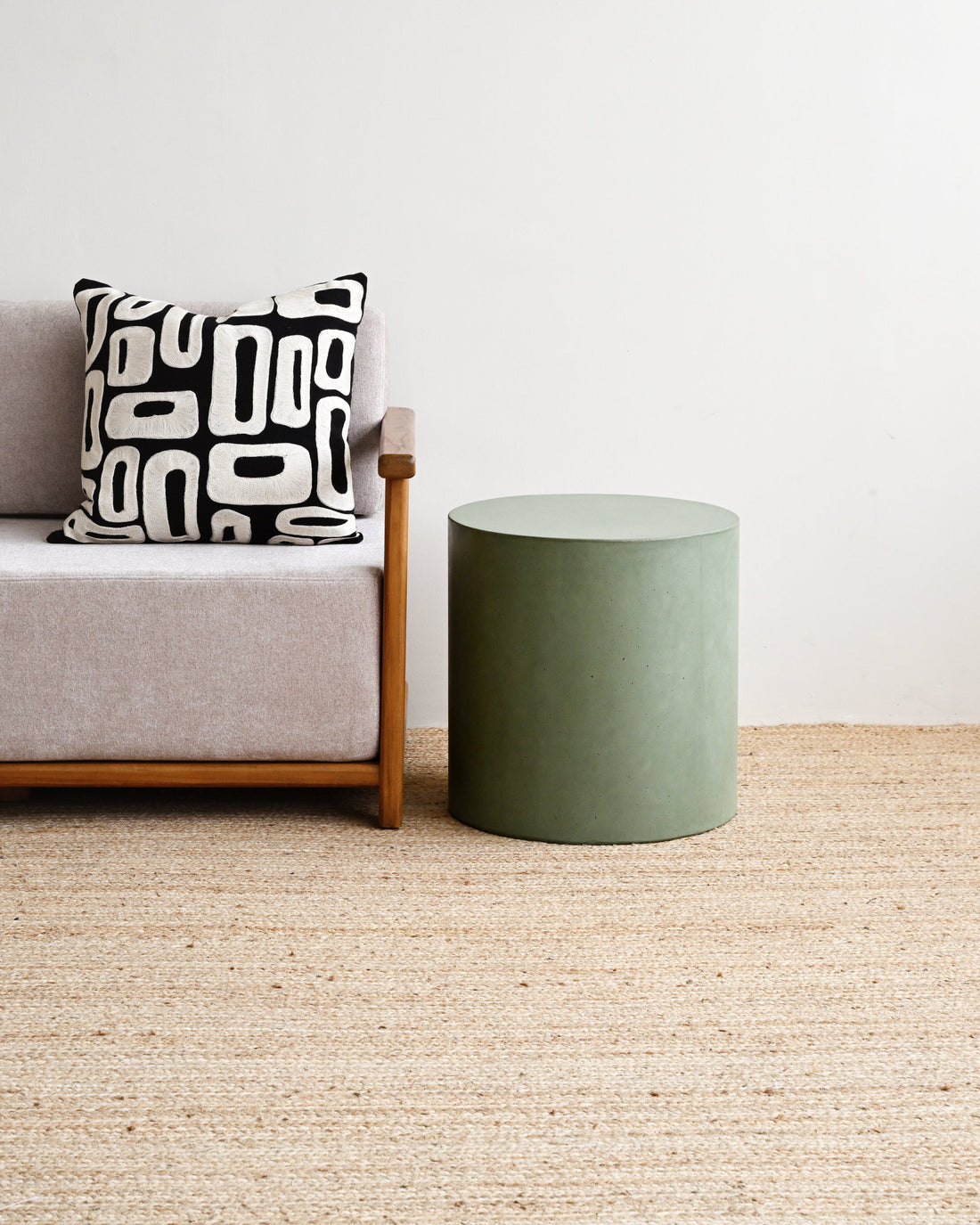 Gray sofa with a black and white patterned pillow next to a green cylindrical side table on a beige carpet.