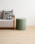 Gray sofa with a black and white patterned pillow next to a green cylindrical side table on a beige carpet.