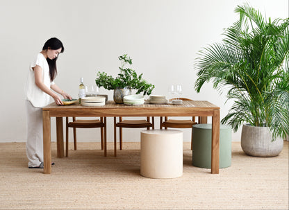Woman arranging items on a wooden dining table in a minimalistic room with plants.
