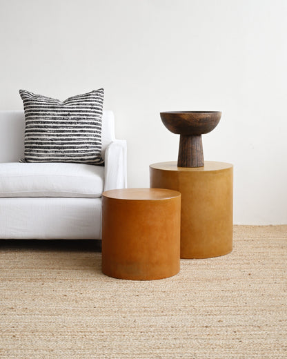 Two brown cylindrical side tables in a living room setting with a white sofa and striped pillow.