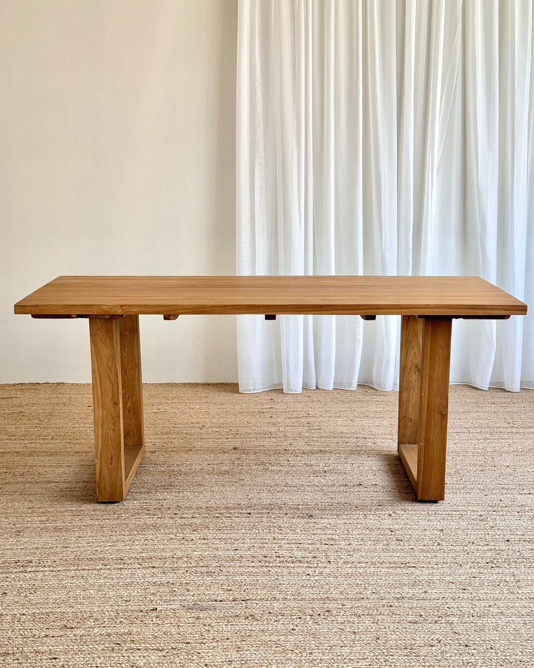 A rectangular wooden dining table with a natural oak finish, featuring thick blocky legs, set on a woven jute rug in front of white curtains.