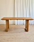 A rectangular wooden dining table with a natural oak finish, featuring thick blocky legs, set on a woven jute rug in front of white curtains.