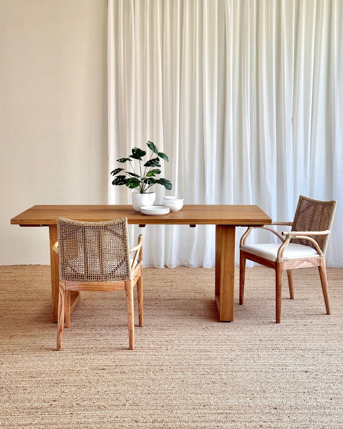 A minimalist dining set featuring a rectangular wooden table and two woven-back chairs with cream cushions, styled on a textured jute rug in front of white curtains.