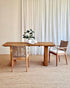 A minimalist dining set featuring a rectangular wooden table and two woven-back chairs with cream cushions, styled on a textured jute rug in front of white curtains.