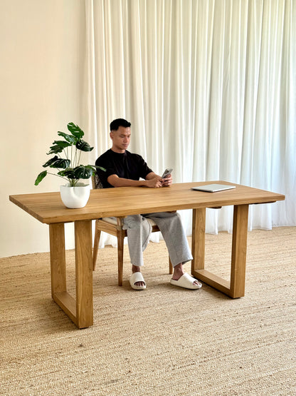 A man sits at a large light oak wooden writing desk in a bright room with cream curtains and a woven rug. The desk has a natural wood grain finish and blocky, geometric legs.