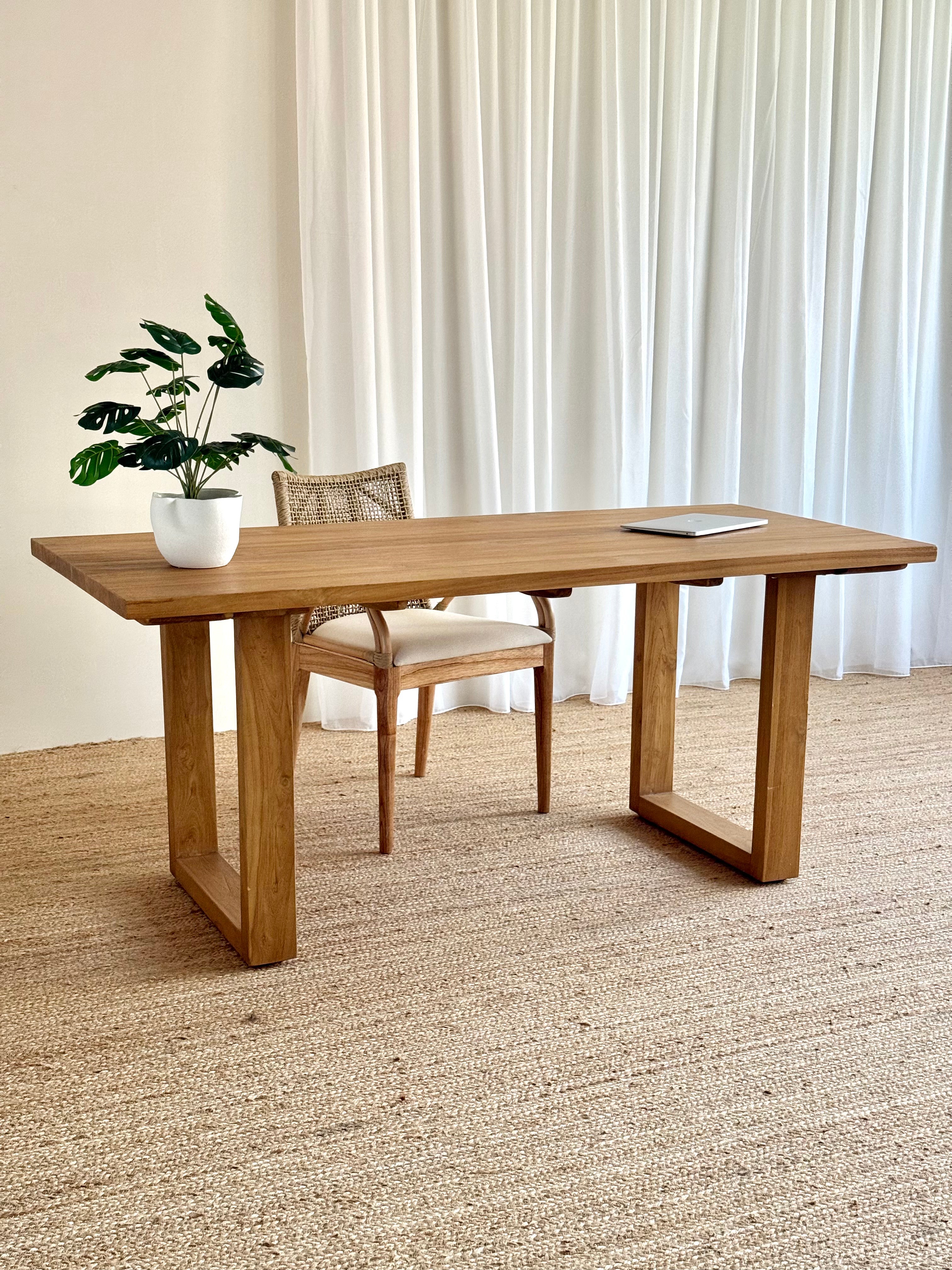 A rectangular wooden writing desk with thick block legs and a matching woven-back chair, styled in a bright room with white curtains, a beige rug, and a potted plant on the tabletop.