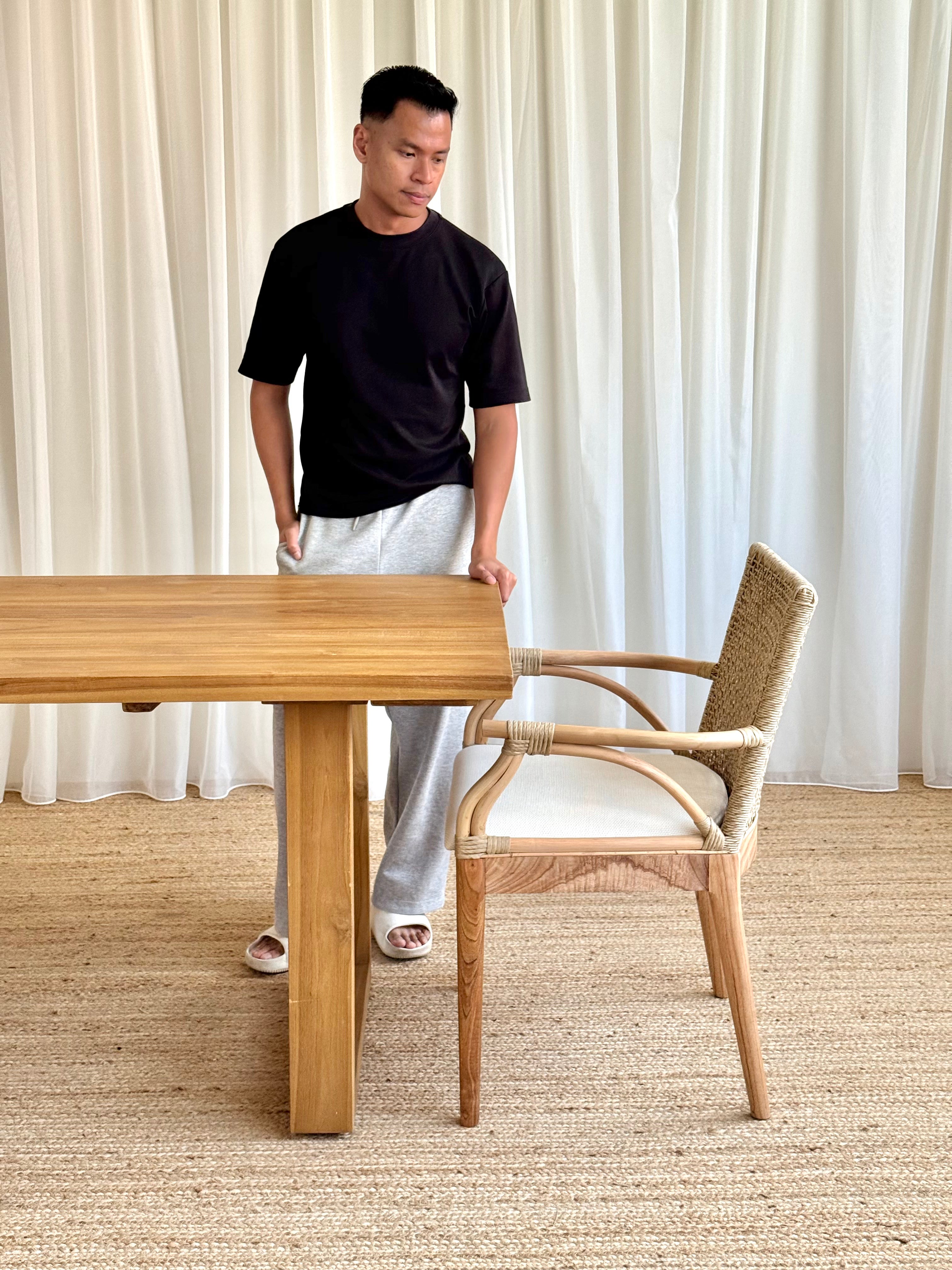 A man in a black t-shirt and white sweatpants stands beside a light wood dining table and a woven rattan armchair in a bright room with white curtains and a beige rug.