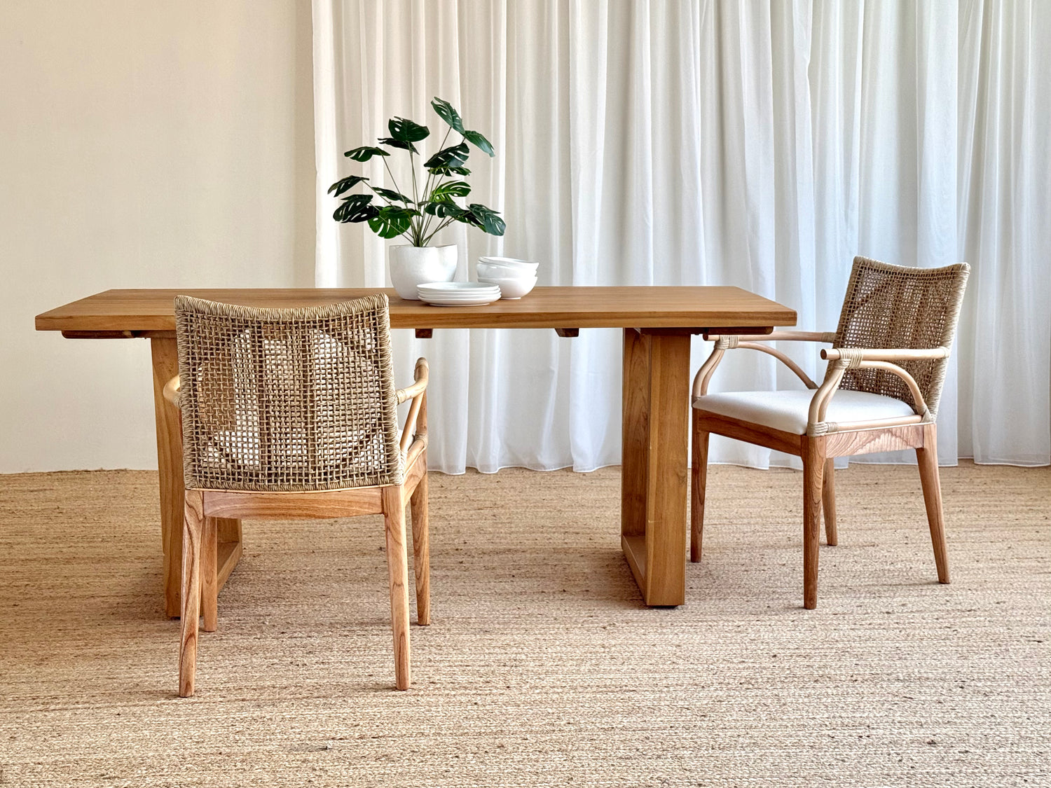 A rectangular wooden dining table with two woven-back chairs in a bright, neutral room with white curtains and a textured rug. A potted plant and small bowls sit on the table.