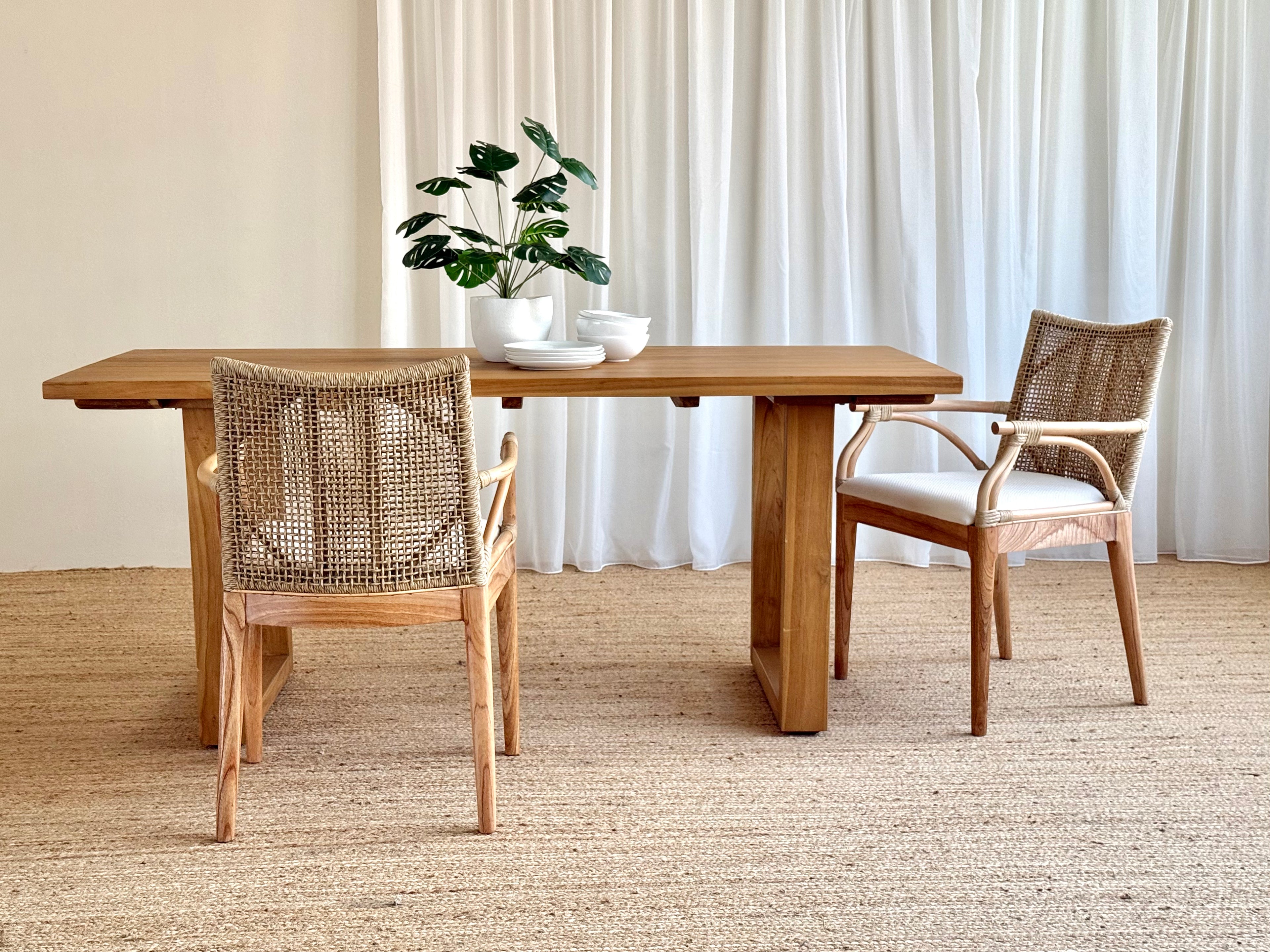 A rectangular wooden dining table with two woven-back chairs in a bright, neutral room with white curtains and a textured rug. A potted plant and small bowls sit on the table.