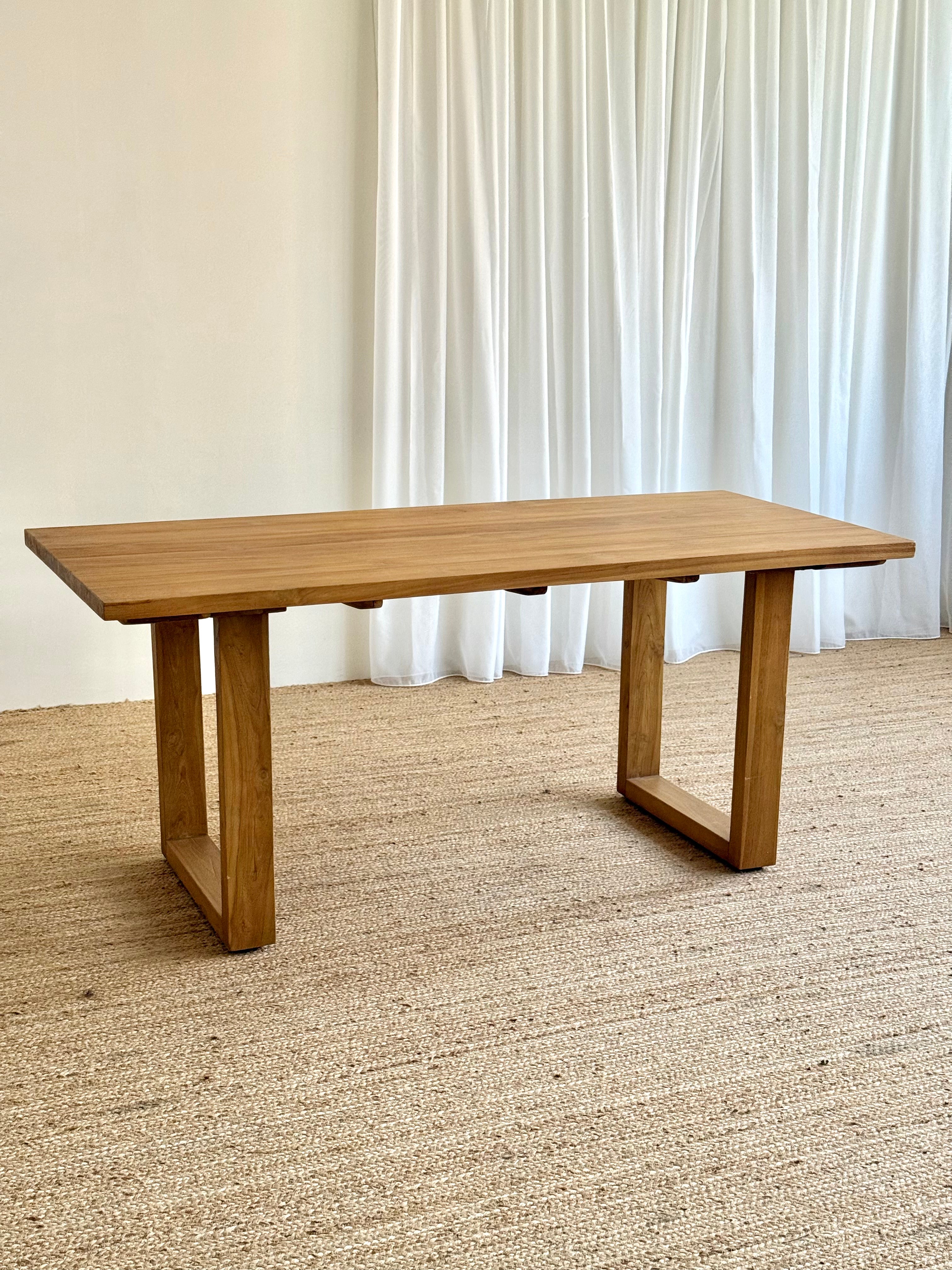 A rectangular wooden dining table with a natural oak finish, featuring thick block-style legs, set on a textured beige jute rug in front of white curtains.
