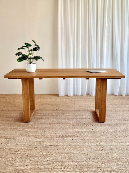 A rectangular wooden writing desk with a warm medium-brown finish and block-style legs, styled in a minimalist room with a potted plant and a laptop on top, set against a neutral beige wall and white curtains.
