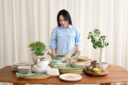 Woman arranging ceramic plates and bowls on a wooden table with plants in the background.