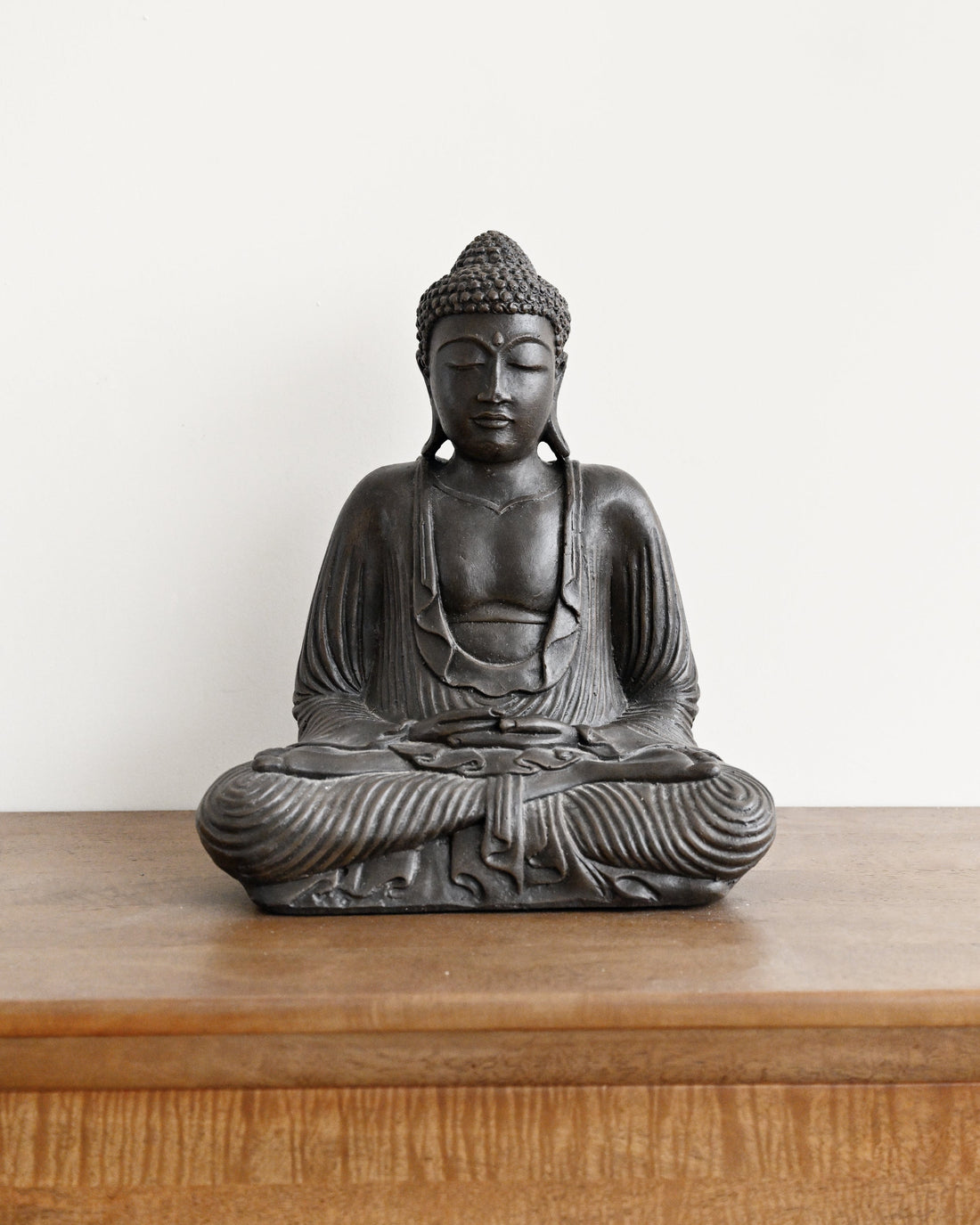A dark gray cast-iron Buddha statue in a seated meditation pose, displayed on a wooden surface against a plain white wall.
