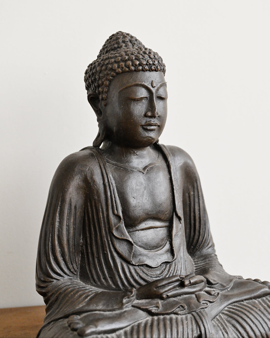 A dark bronze Buddha statue seated in a meditative pose, shown from the chest up against a plain off-white wall.