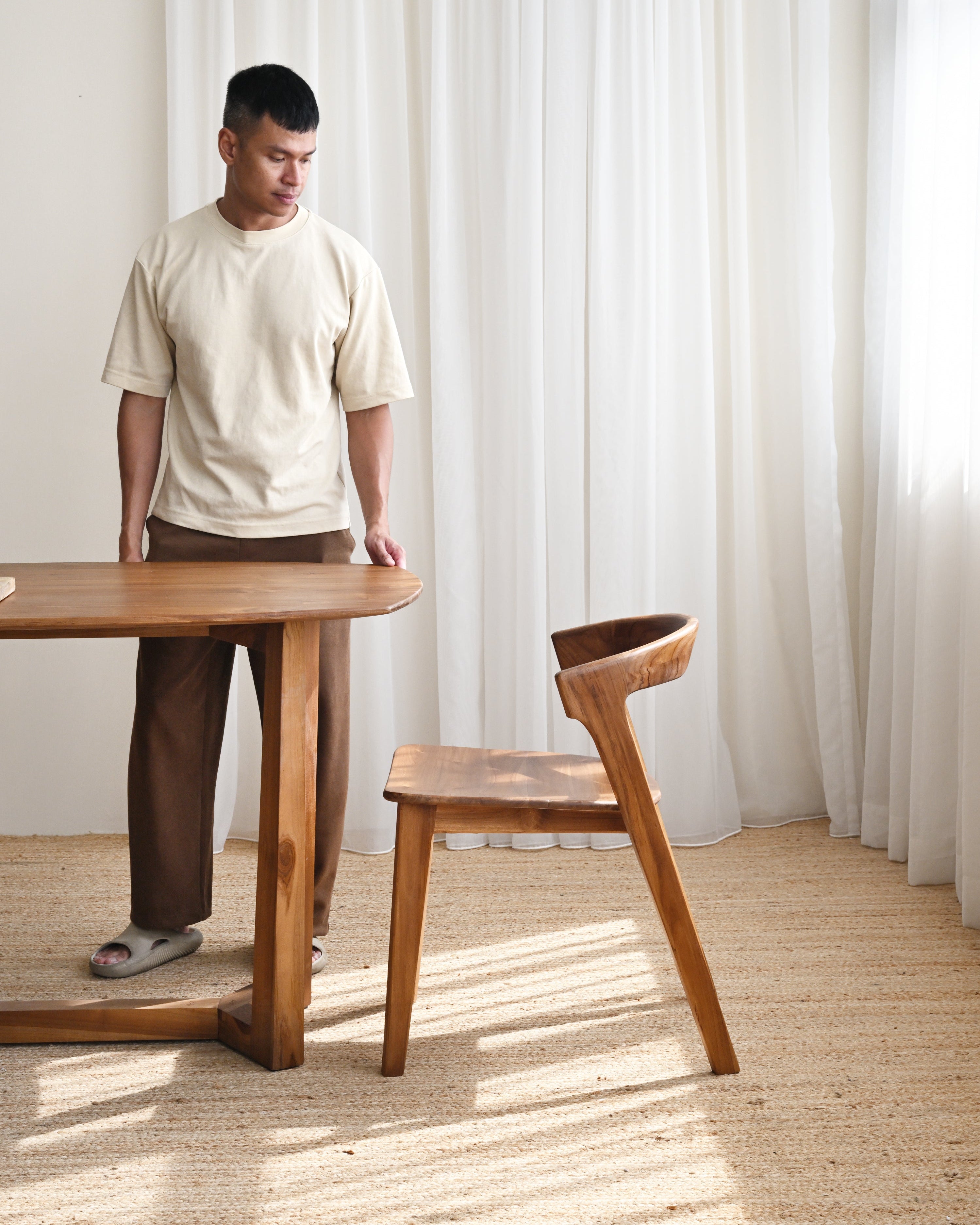 Man standing next to a wooden table and chair in a room with white curtains.