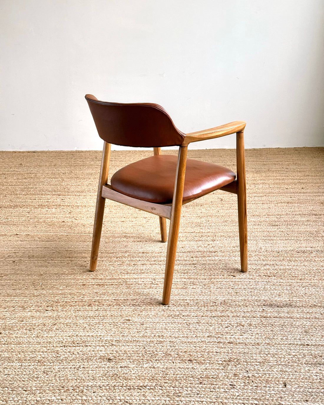 A mid-century modern wooden dining chair with a brown leather upholstered seat and curved backrest, positioned on a textured beige jute rug against a plain off-white wall.