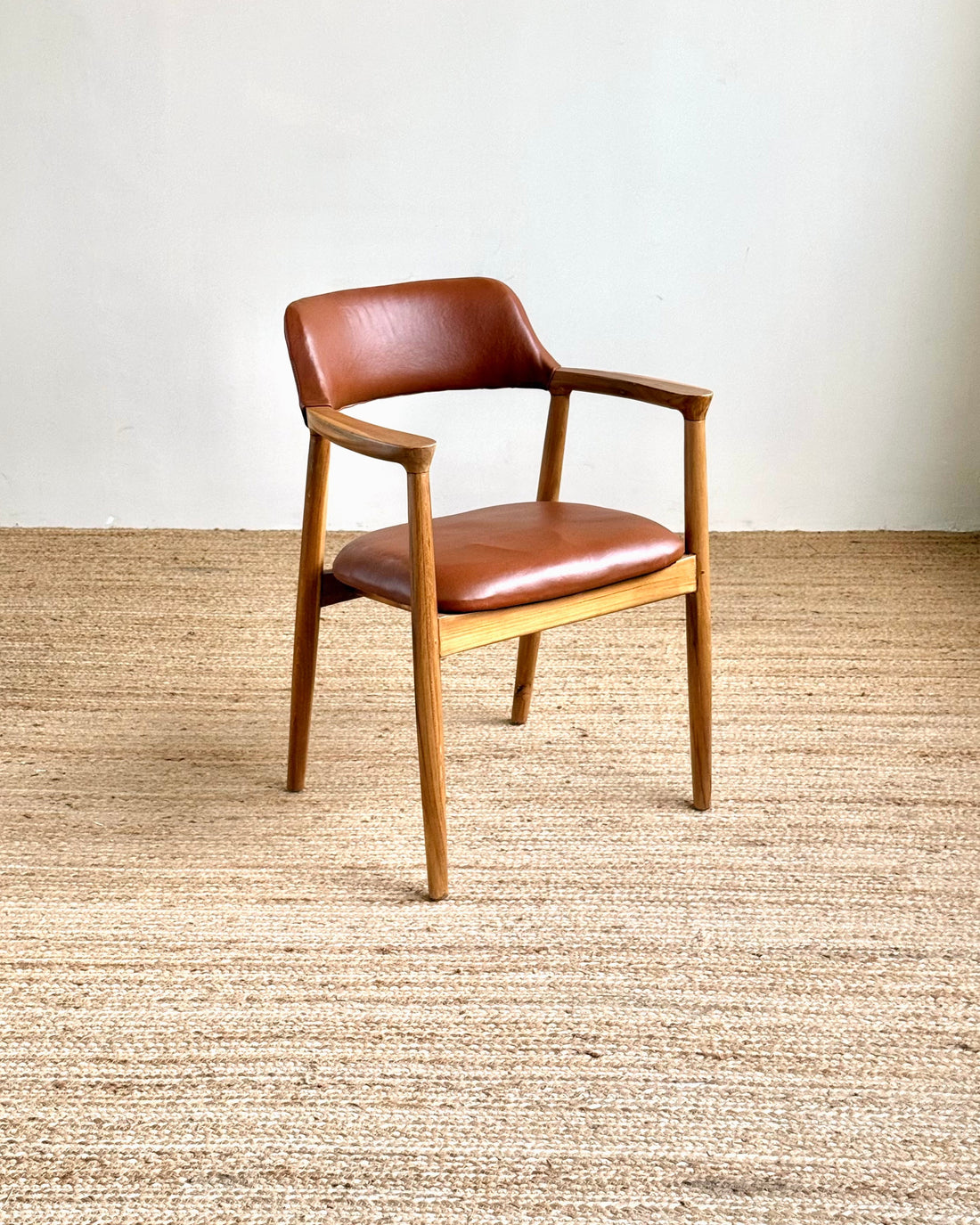 A mid-century modern armchair with a warm brown leather seat and backrest, supported by a light natural wood frame, set on a textured beige woven rug against a plain off-white wall.