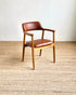 A mid-century modern armchair with a warm brown leather seat and backrest, supported by a light natural wood frame, set on a textured beige woven rug against a plain off-white wall.