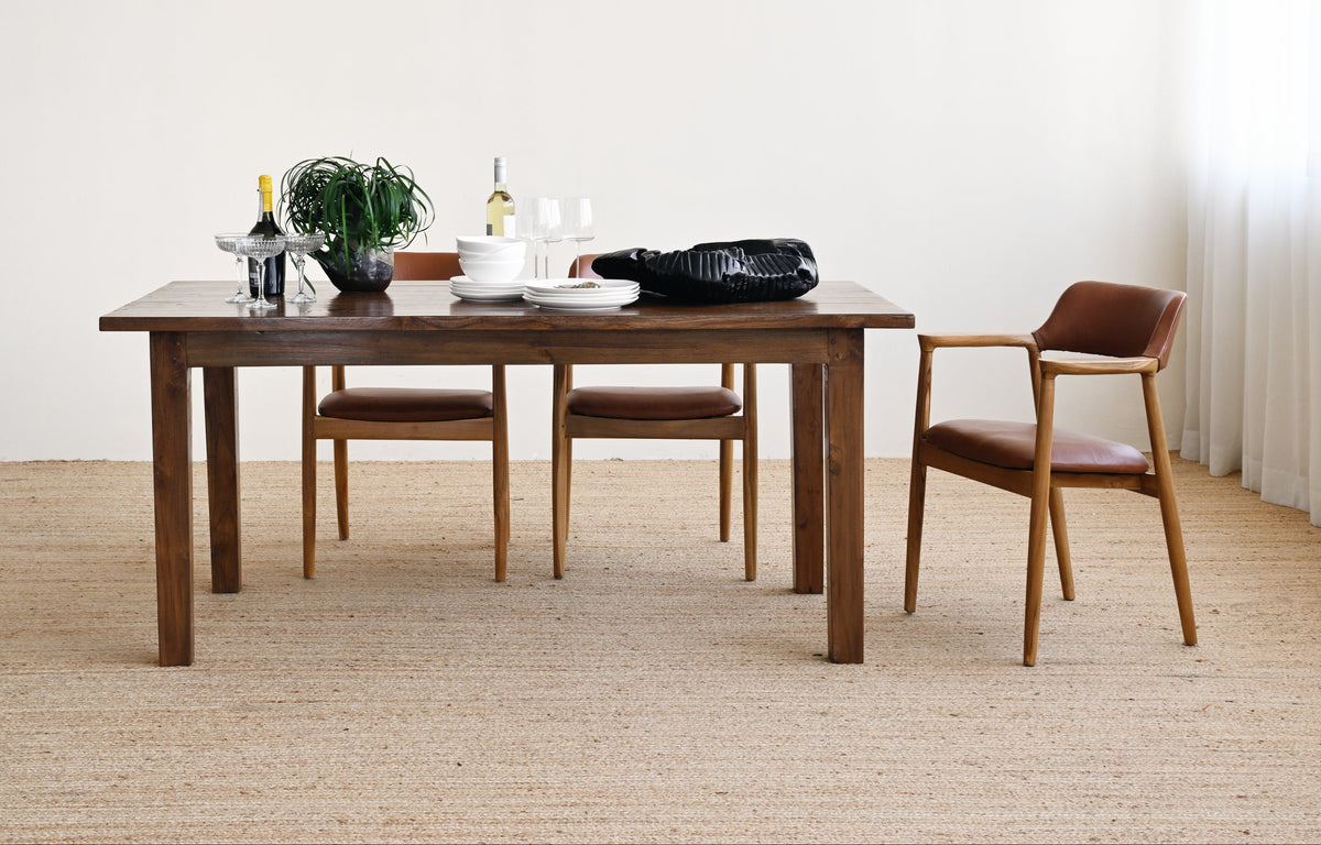 A dark wood dining table with matching chairs in a minimalist room with a light beige woven rug and white walls. The table is styled with a plant, wine bottles, and a black bag.