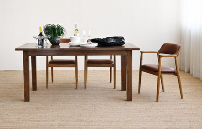 A dark wood dining table with matching chairs in a minimalist room with a light beige woven rug and white walls. The table is styled with a plant, wine bottles, and a black bag.