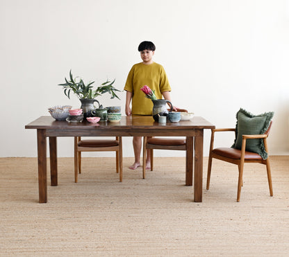 A person wearing a mustard yellow t-shirt and shorts stands behind a dark wood dining table set with bowls, a plant, and chairs in a minimalist room with a light beige wall and woven floor.