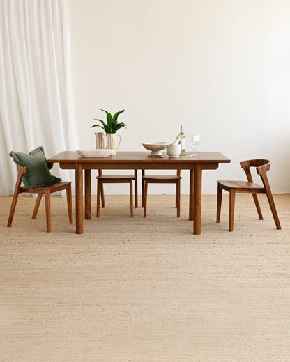 A mid-century modern wooden dining table set with four matching wooden chairs, styled with a small plant and ceramic dishes on a light beige woven rug against a neutral cream wall.
