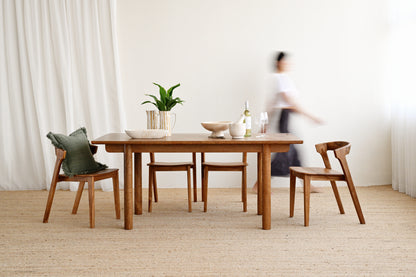 A wooden dining table with four matching wooden chairs in a bright, minimalist room with white curtains and a light beige rug. A green pillow rests on one chair, and a person is blurred in the background near the table.