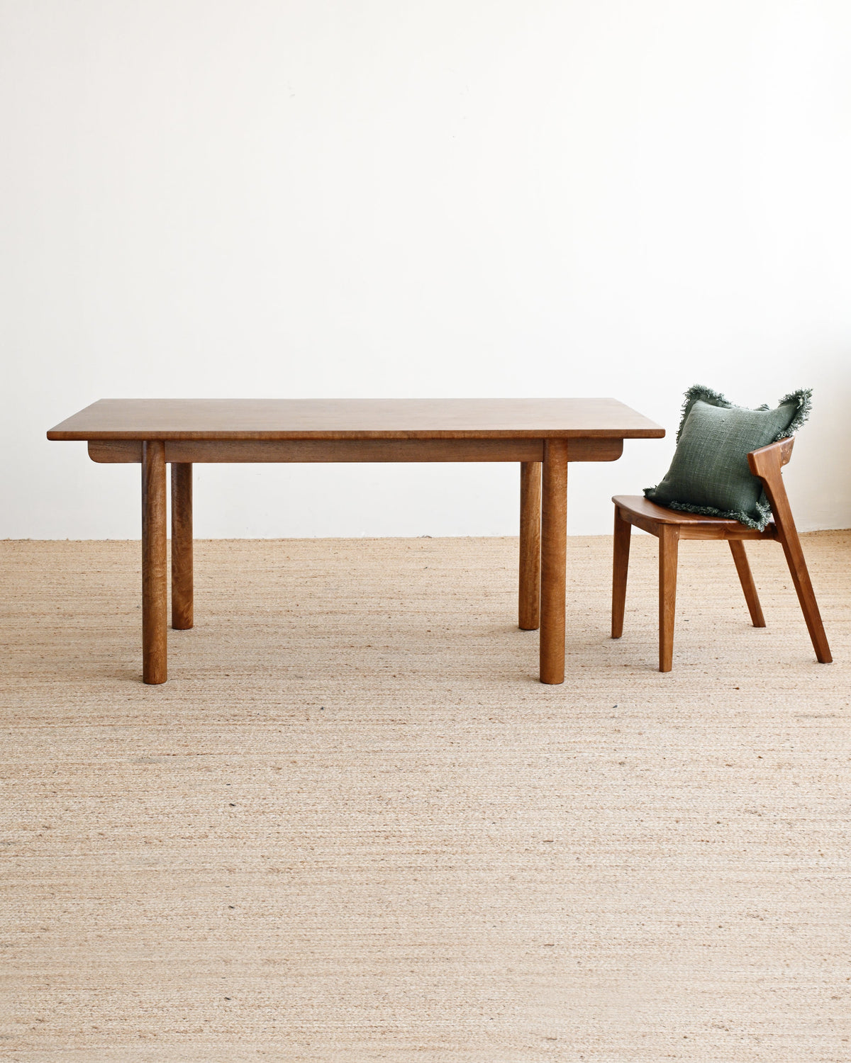 A minimalist wooden dining table with a matching wooden chair holding a green pillow, set on a light beige woven rug against a plain white wall.